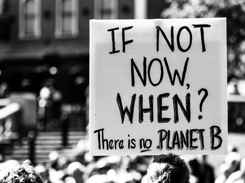 Black and white photograph of a climate protest. Someone is holding up a sign saying "If not now, when? There is not PLANET B"
