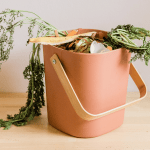Indoor brick-orange food waste bin. Leafy greens of carrots poking out of the top. Sitting on a wooden surface set against a plain grey wall.
