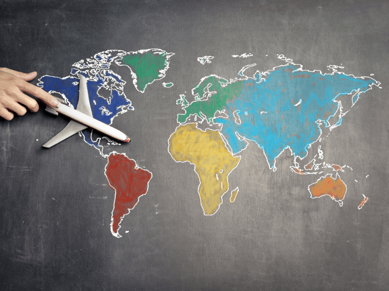 Map of the world drawn in white on a black chalkboard. Each continent shaded in a different chalk colour: red, dark blue, light blue, yellow, orange, green. A white hand coming in on the left over North America holding a toy plane pointing across the map.