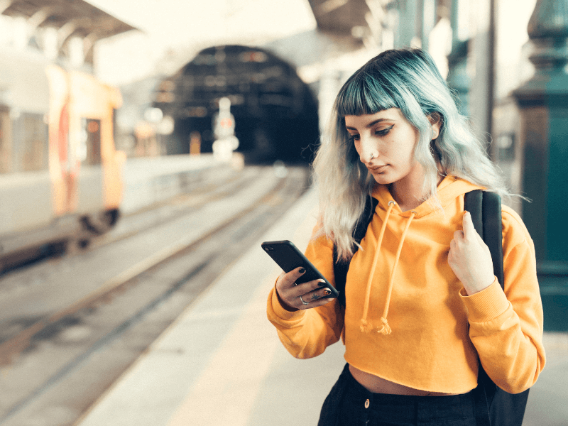 A girl standing at a train station holding out her phone. She is wearing a yellow jumper, a black rucksack, and has blue fringed hair.