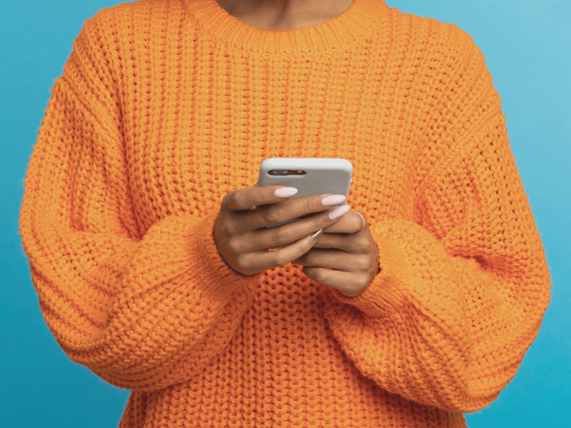 Woman in orange knitted jumper standing in front of a blue background holding her phone.